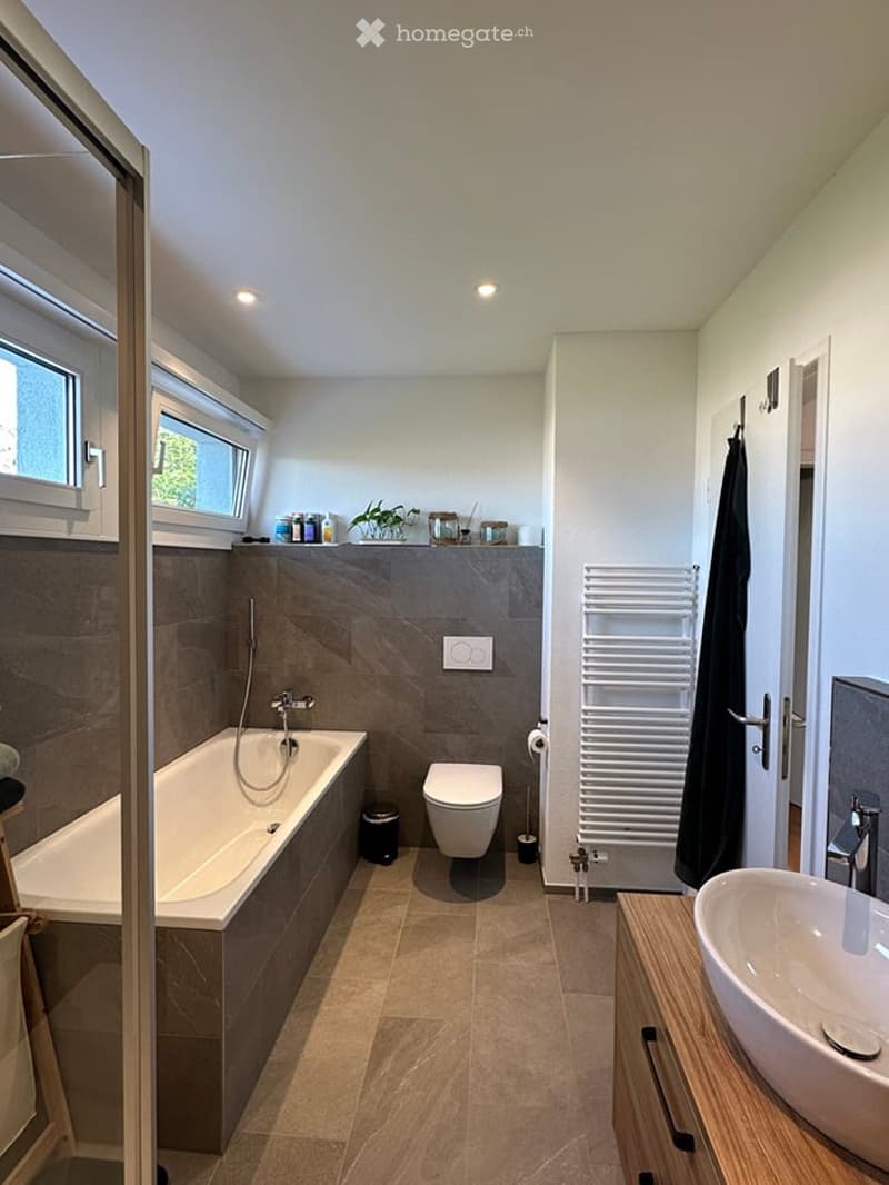 Modern bathroom with a bathtub, toilet, sink, and radiator. Features include a large window, tiled walls, and a sleek design.