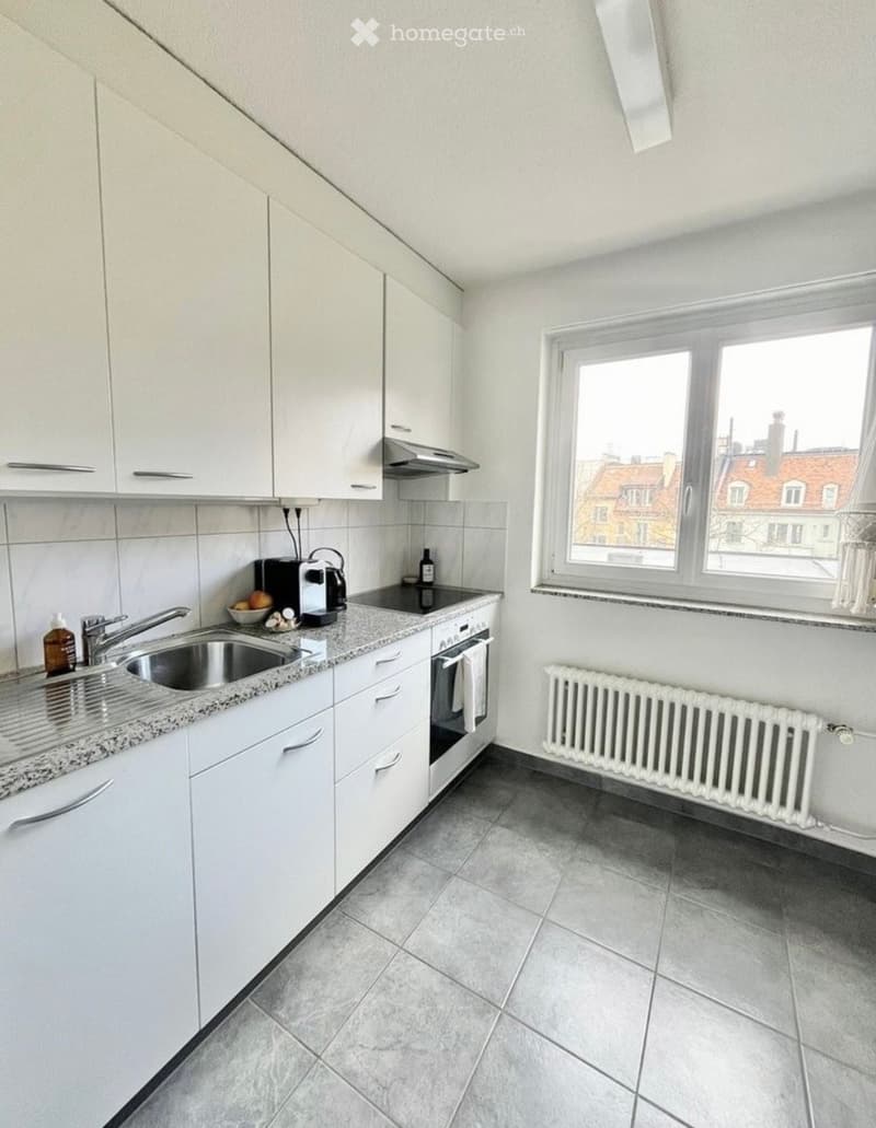 Modern kitchen with white cabinets, granite countertop, stainless steel sink, and built-in appliances. Features a window with a city view and a radiator below.