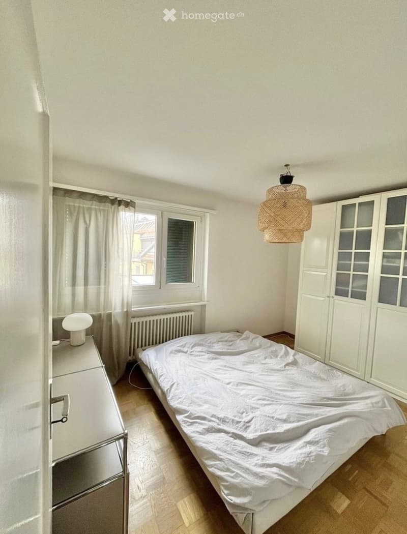 Minimalist bedroom with a single bed, white wardrobe, hanging woven lamp, and a small nightstand with a lamp.