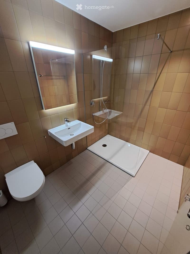 Modern bathroom with beige tiles, a toilet, a sink, a shower, and a large mirror with built-in lighting.