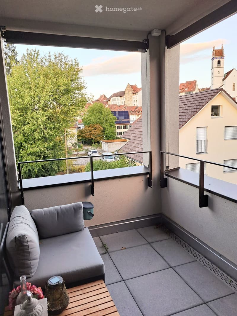 Balcony with a grey sofa, small wooden table, vase with flowers, and a scenic city view.