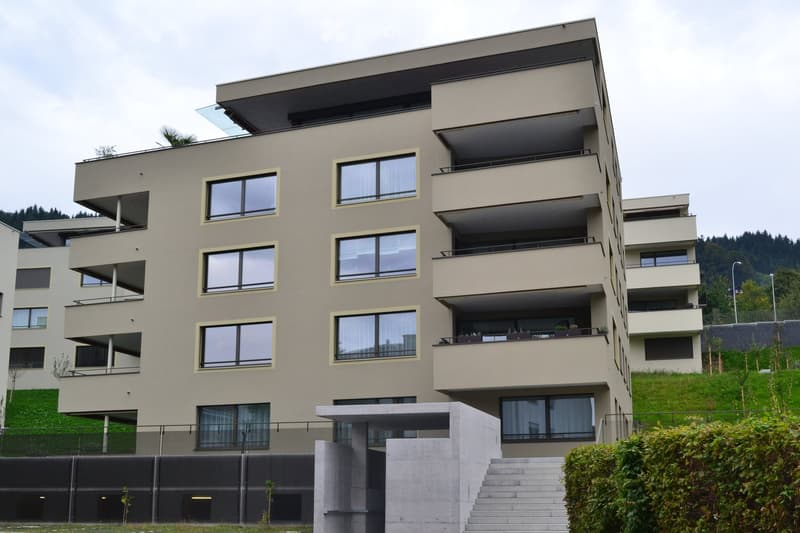 Modern multi-story residential building with large windows, balconies, and a flat roof, surrounded by greenery and a staircase leading to the entrance.