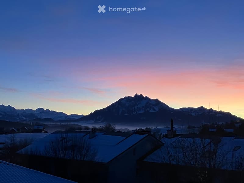 A serene mountain landscape at dawn, with snow-covered roofs in the foreground and a range of snow-capped mountains in the background.
