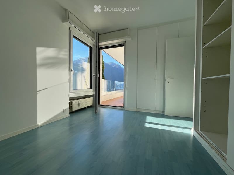 Empty room with light blue wooden flooring, white walls, large windows, and a sliding glass door leading to a balcony with a scenic mountain view.