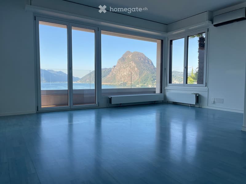 Empty room with large windows offering a scenic mountain and lake view, light-colored wooden flooring, and white walls.