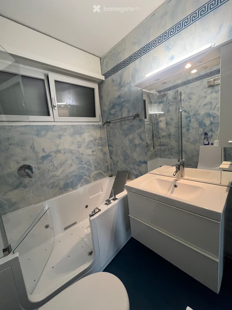 Modern bathroom with a white bathtub, double sink vanity, toilet, and a glass-enclosed shower. The walls are covered in blue marble tiles with a decorative border, and there is a window with blinds above the bathtub.