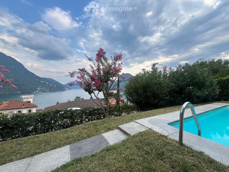 Garden area with a swimming pool, lush greenery, and a scenic view of a lake and mountains in the background.