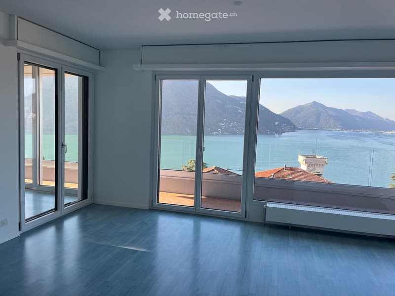 Bright, empty room with large windows and sliding doors opening to a balcony, overlooking a scenic lake and mountains.