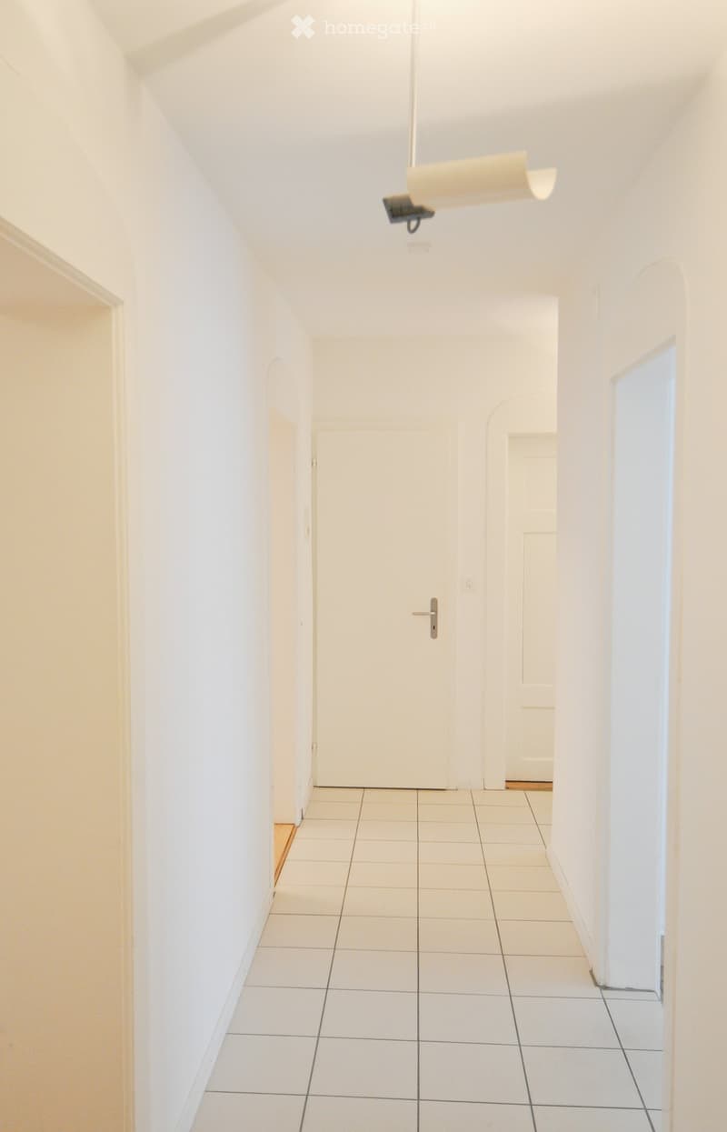A clean, white-walled hallway with tiled flooring, featuring a closed door at the end and a modern light fixture hanging from the ceiling.