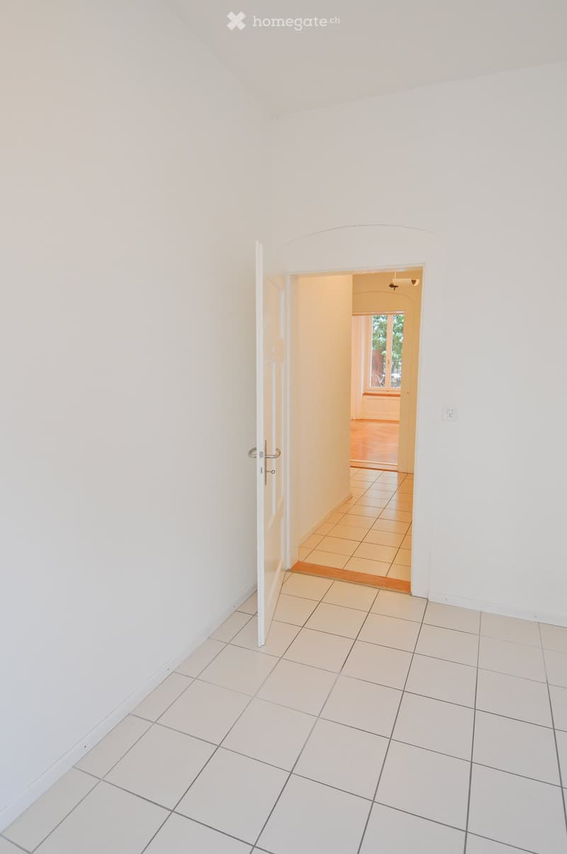 Empty hallway with white tiled flooring and white walls, featuring an open doorway leading to another room with wooden flooring and a window.
