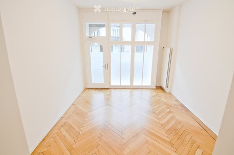 Empty room with light wooden herringbone flooring, white walls, large glass doors leading outside, and a radiator on the right wall.