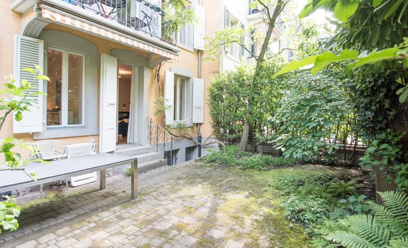 Garden area with a table and chairs, surrounded by greenery and a building entrance.