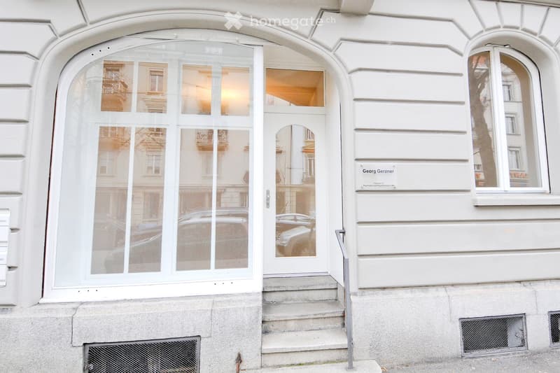 Facade of a white building with large arched windows and a glass door entrance. The entrance has a small staircase leading up to it, and a nameplate is visible on the wall to the right.