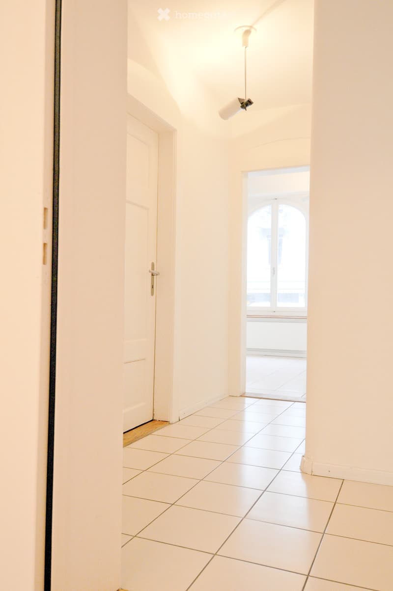Empty hallway with white tiled floor, white walls, and a single door on the left leading to another room. Natural light streams in from an arched window at the end of the hallway.