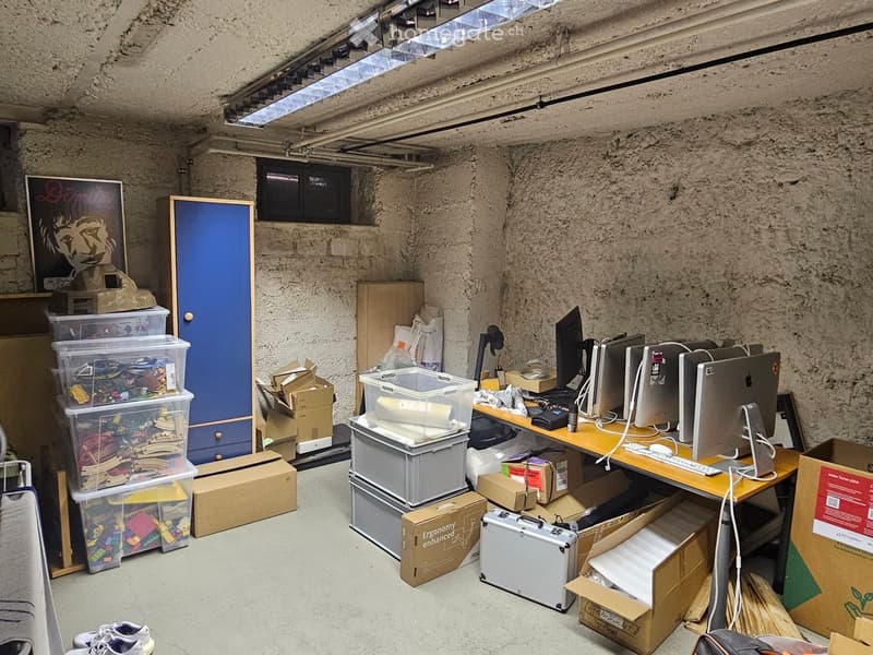 A cluttered room with a desk setup, multiple computer screens, a keyboard, mouse, storage bins, and various boxes scattered around.