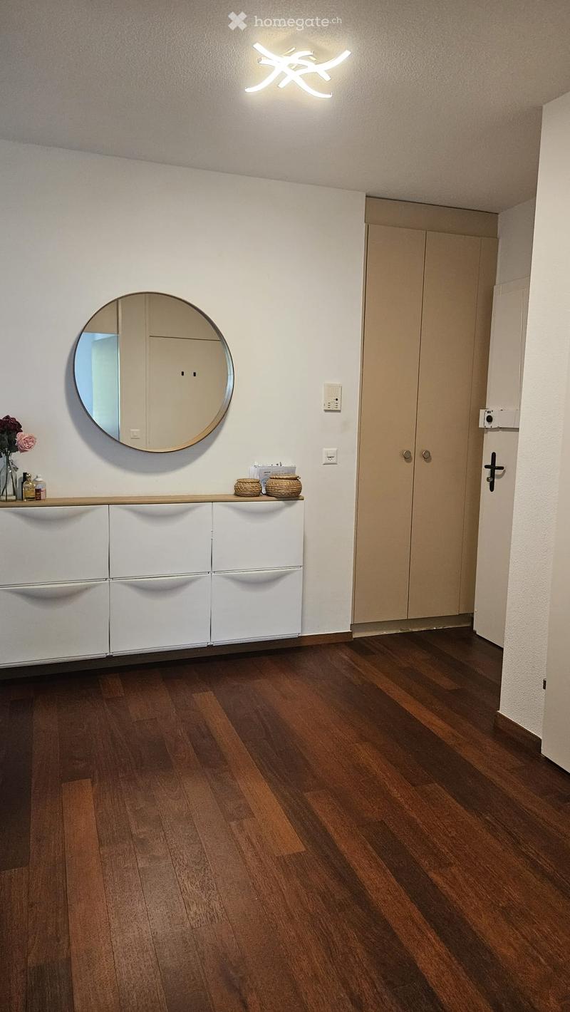 Hallway with wooden flooring, white cabinetry, round mirror, decorative items on the cabinet, and a closed door.