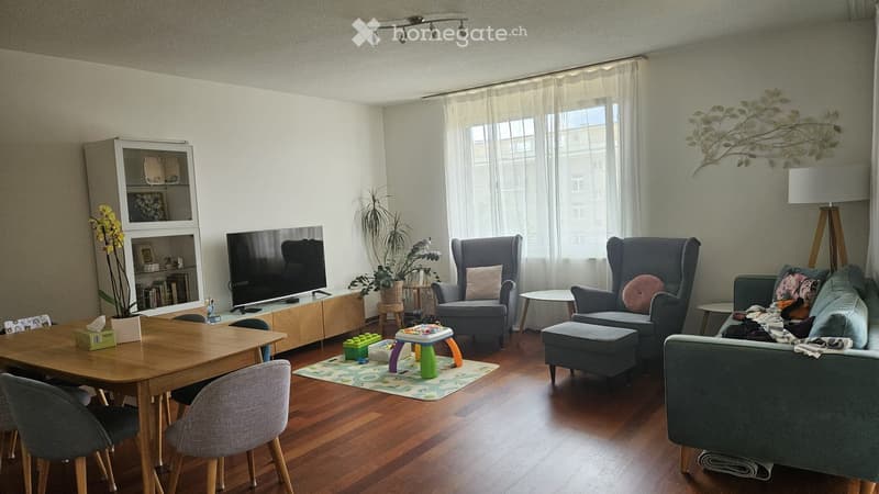 A well-lit living room with wooden flooring, a large window with white curtains, a grey sofa, armchairs, a coffee table, a dining table with chairs, a television, a bookshelf, and decorative plants.