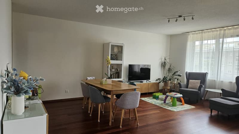 A modern living room with wooden flooring, a large window with sheer curtains, a dining table with chairs, a sofa, a television, and decorative plants.