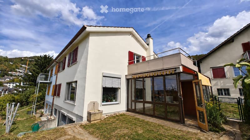 Two-story white house with red shutters, large windows, and a balcony. The house features a modern design with a covered entrance and a small garden area.
