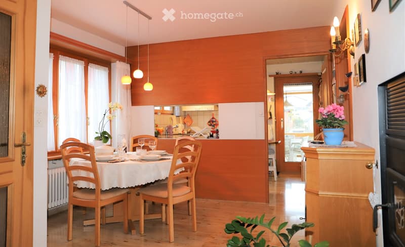 Dining room with a wooden dining table, chairs, and a view into a kitchen area. The room features warm orange walls, wooden flooring, and hanging pendant lights.