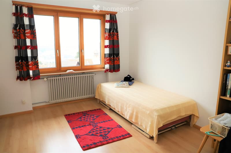 Cozy bedroom with wooden flooring, a single bed with a light-colored bedspread, a red rug, a wooden shelf with books, and a radiator beneath a large window with patterned curtains.