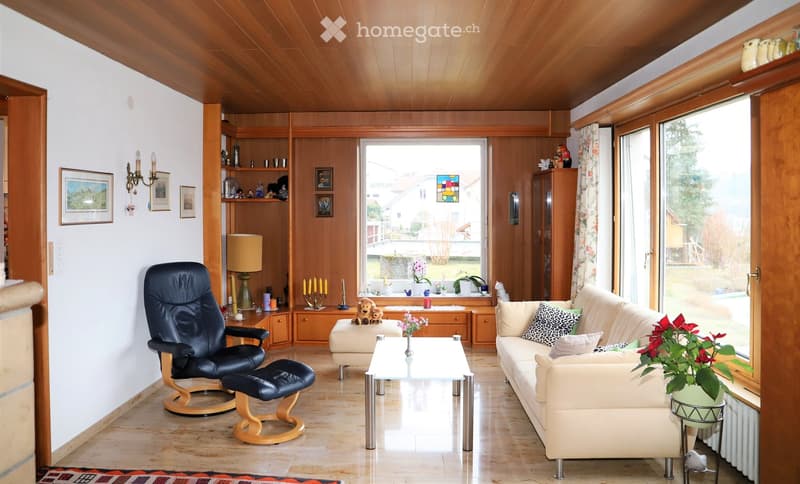 Bright living room with wooden ceiling and walls, large windows, cream sofa, black leather chair, glass coffee table, table lamp, and decorative items on shelves.