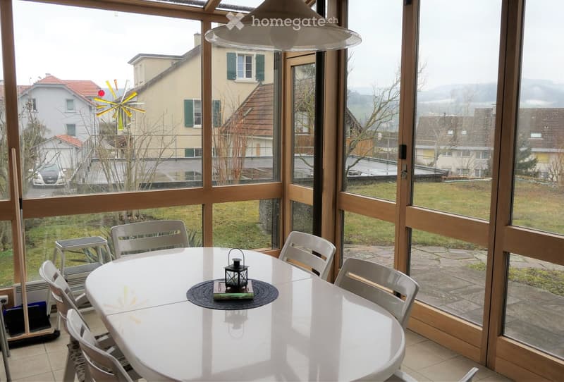 Bright room with large windows overlooking a garden, featuring a round white table with chairs, a lantern, and a book on the table.