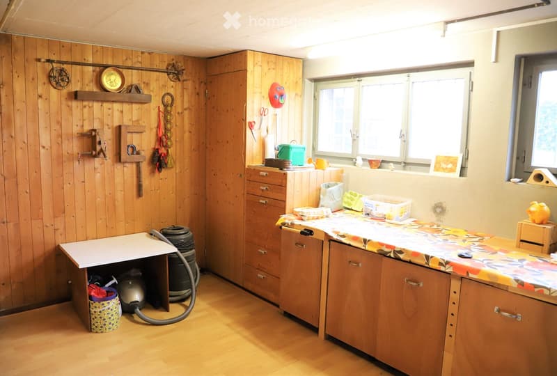 A well-lit room with wooden walls and flooring, featuring a cabinet, a table with a floral cover, a vacuum cleaner, and various decorative items.