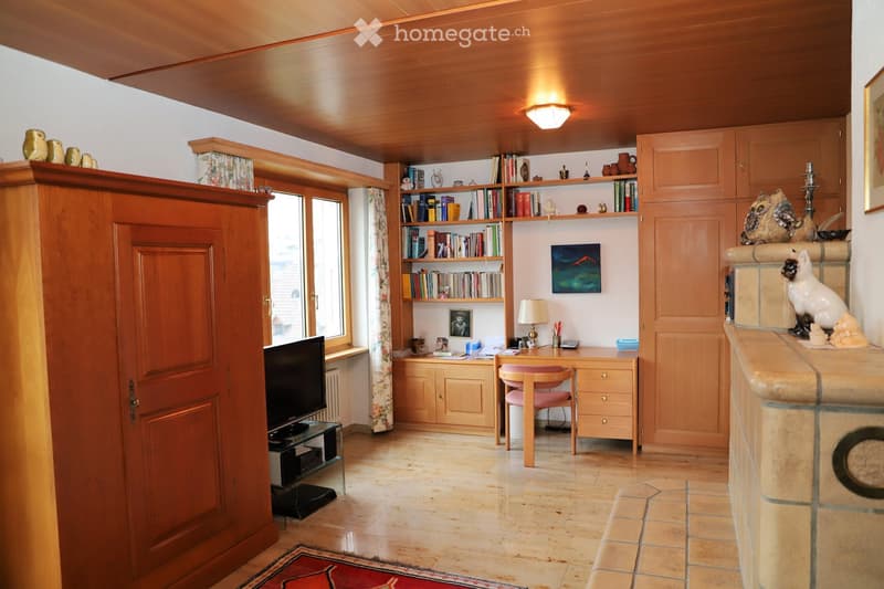 A cozy, well-furnished room with wooden furniture, a desk, bookshelf, and a comfortable chair. The room features a warm wooden ceiling, a fireplace with decorative items, and a large window with curtains.