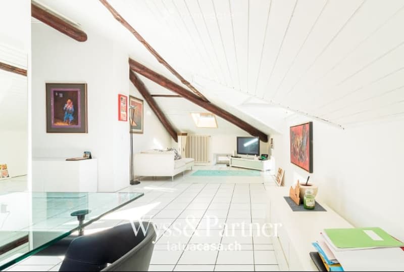 Bright attic living space with exposed wooden beams, white walls, tiled floor, and modern furniture including a sofa, coffee table, chair, and television.