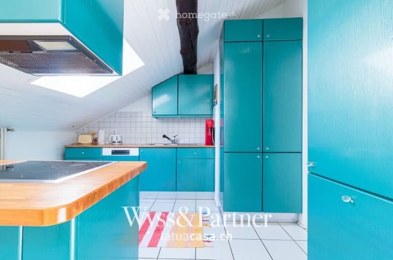 Modern kitchen with teal cabinets, white tiled walls, and a wooden countertop. Features include a sink, faucet, refrigerator, dishwasher, and coffee maker.