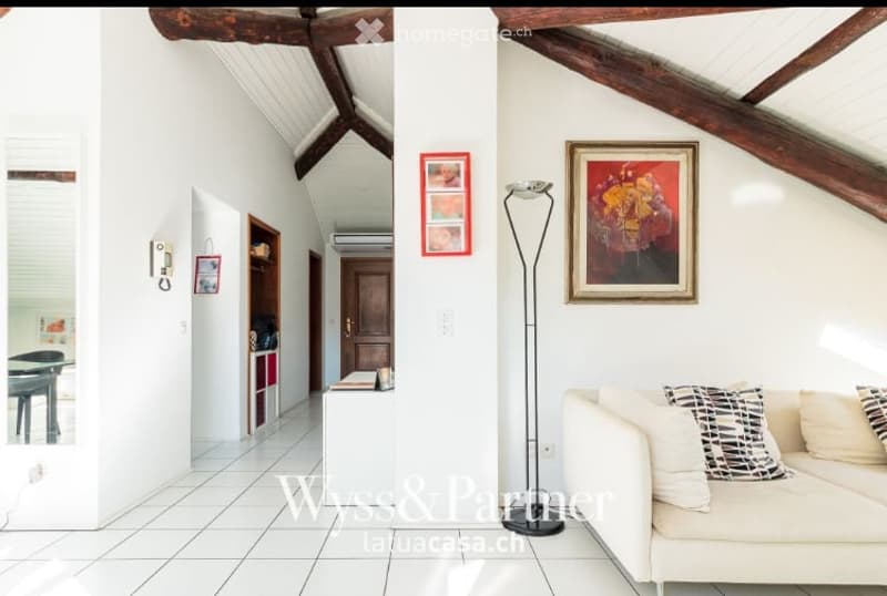 Bright living room with white walls, wooden beams, and a high ceiling. Features a white sofa, floor lamp, small table, and picture frames on the walls.