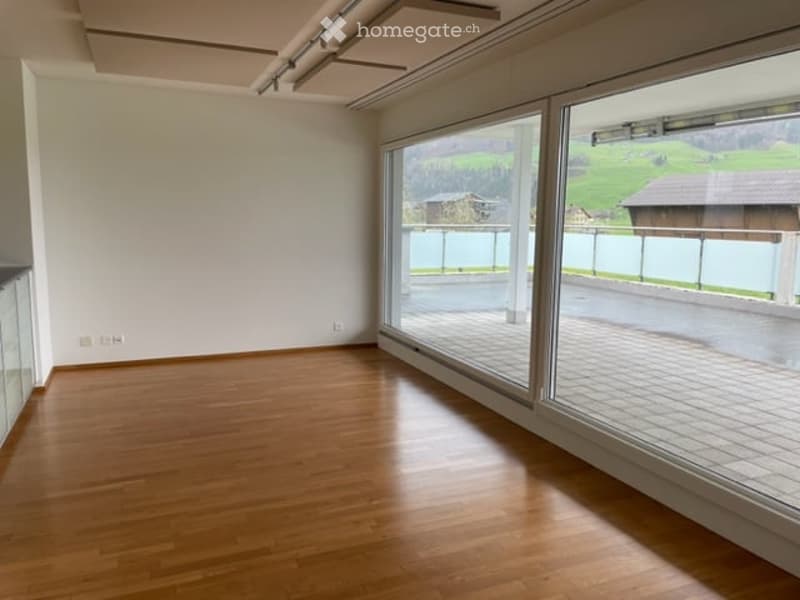 Empty room with wooden flooring, large windows offering a scenic view of green hills and a wooden house.