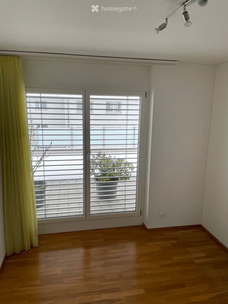 Empty room with wooden flooring, large window with blinds, and a view of a balcony with potted plants.