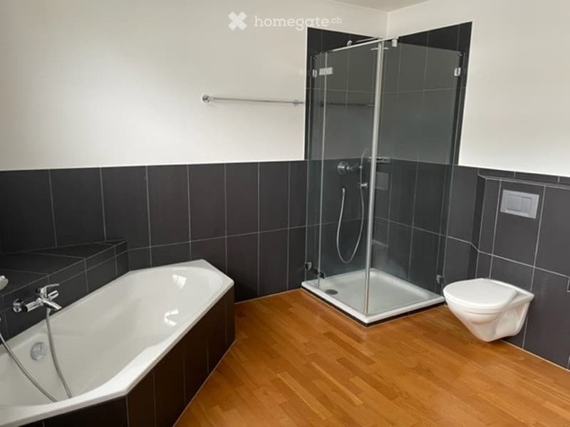 Modern bathroom with a bathtub, toilet, and glass-enclosed shower stall. The walls are tiled in black and white, and the floor is wooden.