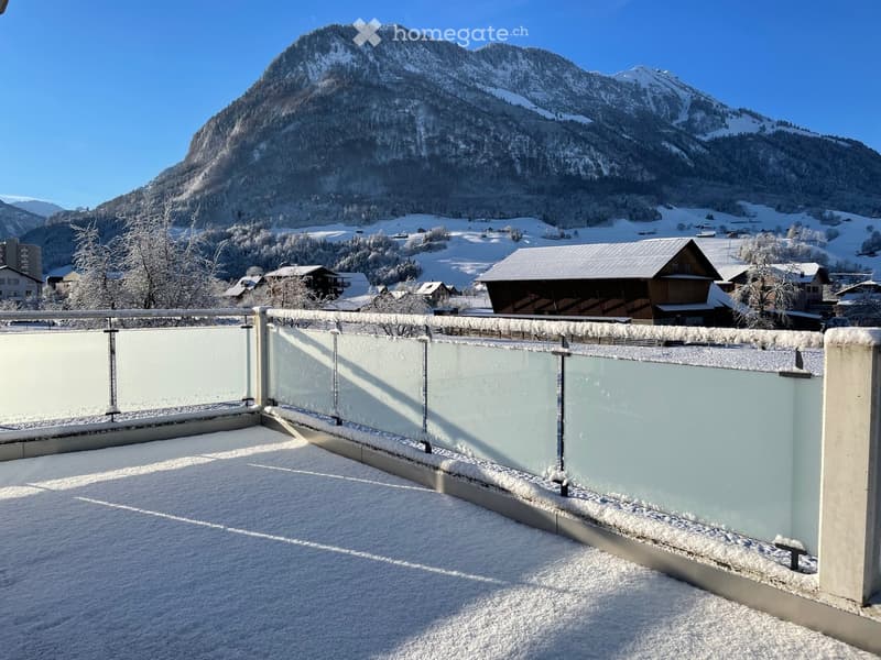 Snow-covered balcony with glass railing, overlooking a snowy mountain landscape and village houses.
