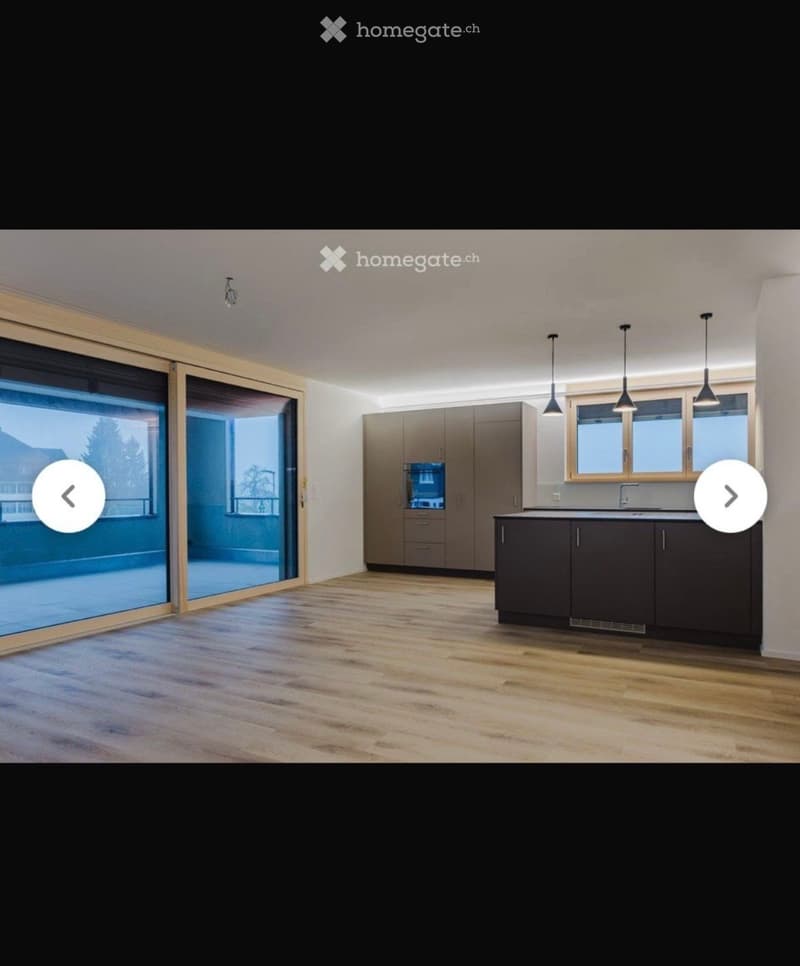 Modern kitchen with light wood flooring, grey cabinets, and a large window overlooking a balcony. The space is clean and well-lit with pendant lights above the counter.