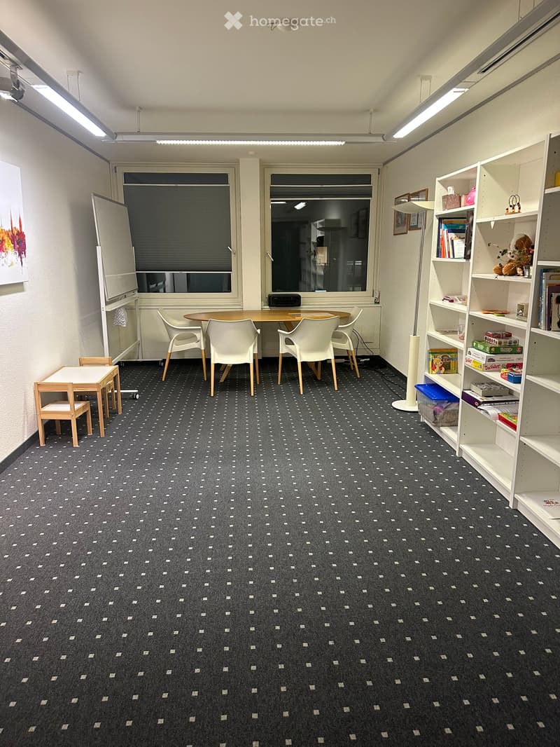 A furnished room with a patterned carpet, a round table with four chairs, a small wooden table with two chairs, a whiteboard, a tall floor lamp, and a white shelving unit with books and decorative items.