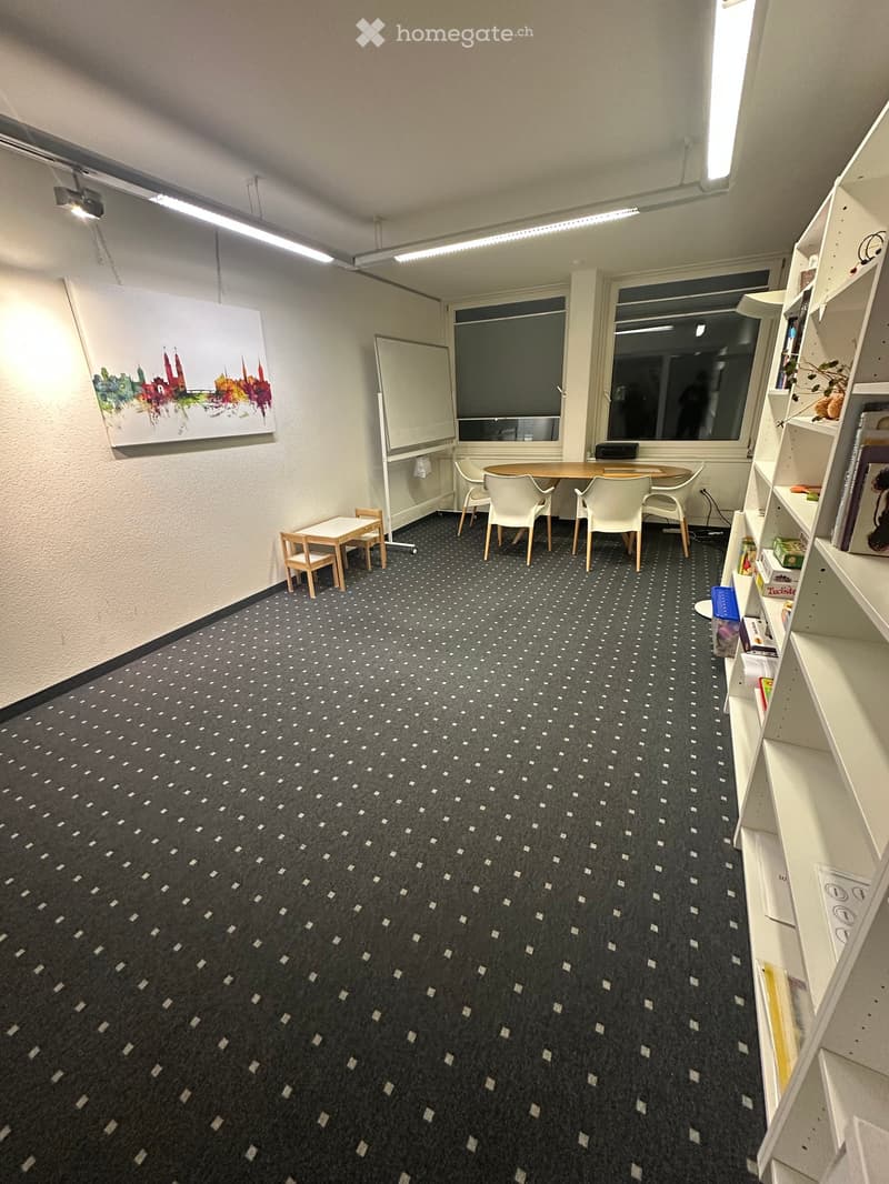 A small, furnished room with a patterned carpet, a large window, a round table with four chairs, a small stool, a whiteboard, and a bookshelf filled with books and decorative items.