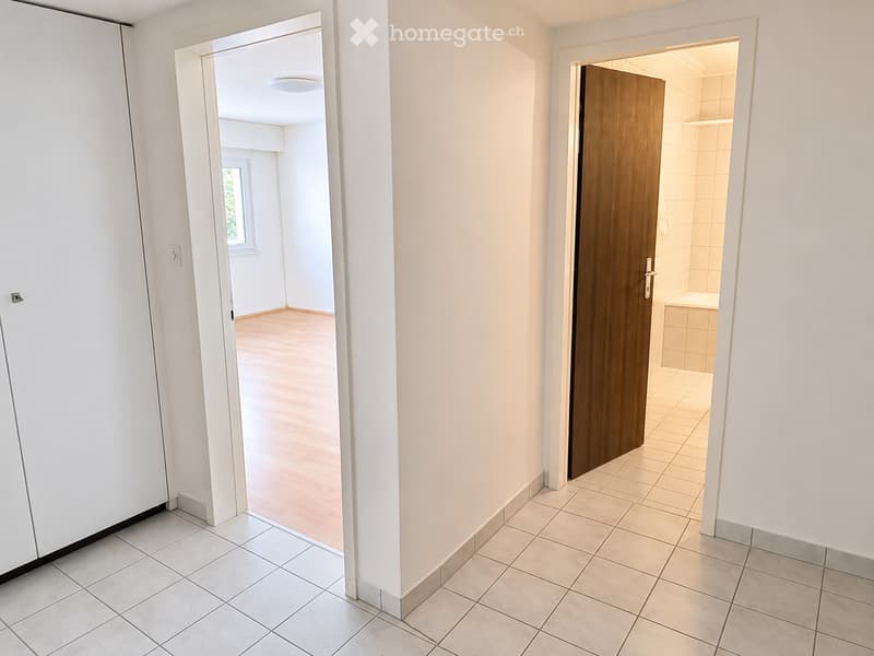 Empty hallway with tiled floor, white walls, and two open doors leading to a room and a bathroom.