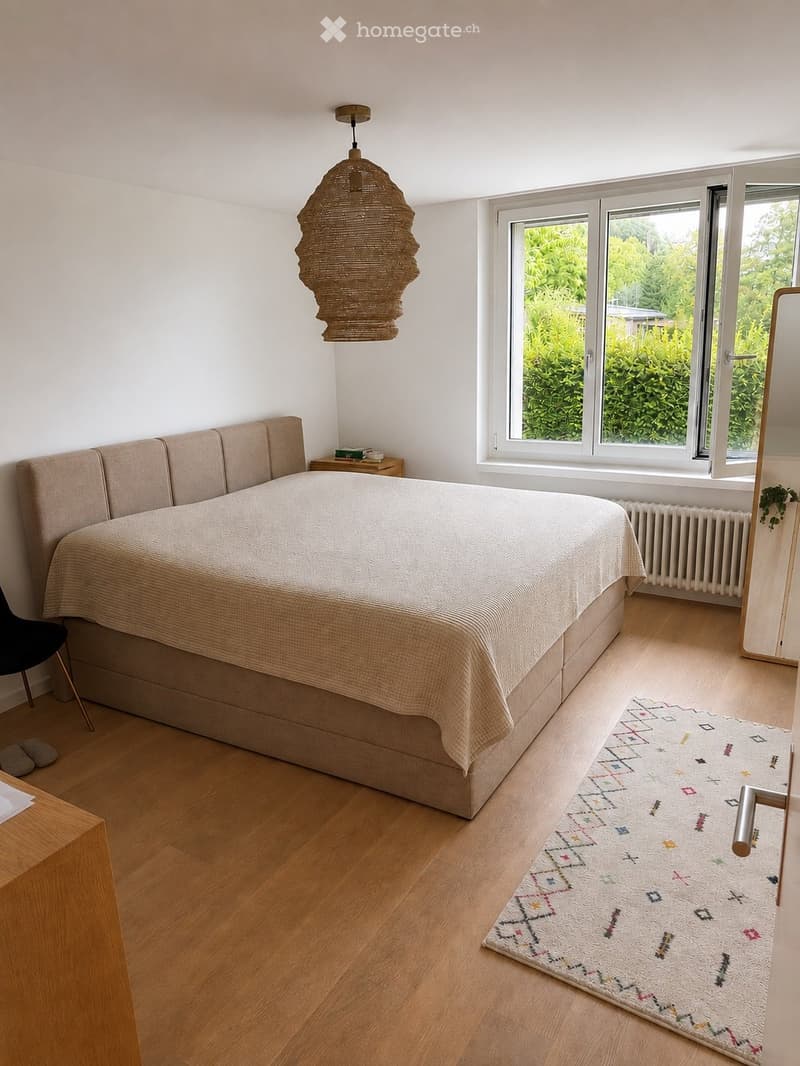 A modern bedroom with a large bed, a wooden side table, a hanging lamp, a patterned rug, and a black chair near the bed.