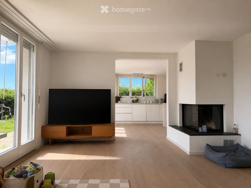 Bright living room with wooden flooring, large television on a wooden stand, fireplace with a black surround, and an open kitchen area with white cabinets. A dog bed is placed near the fireplace, and a wicker basket with toys is visible in the foreground.