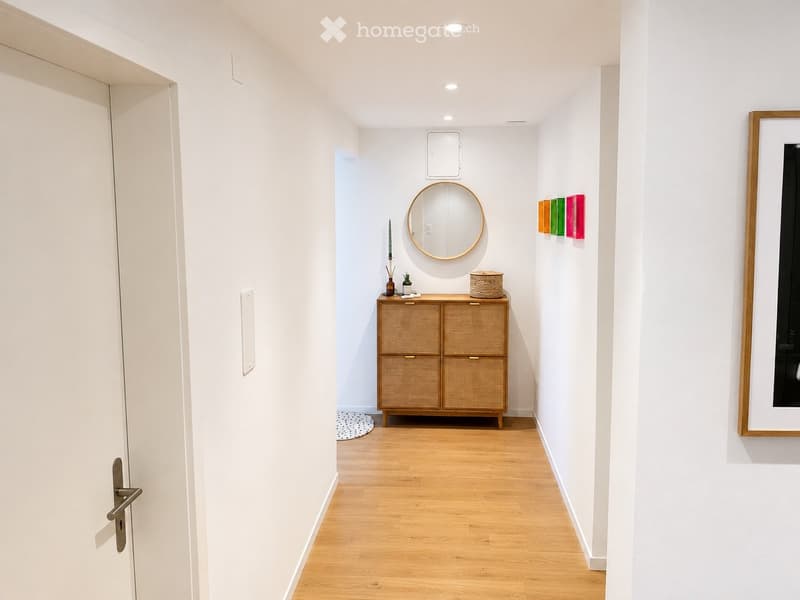 A modern hallway with light wooden flooring, white walls, and recessed ceiling lighting. Features a wicker cabinet with drawers, a round mirror, and colorful wall art.