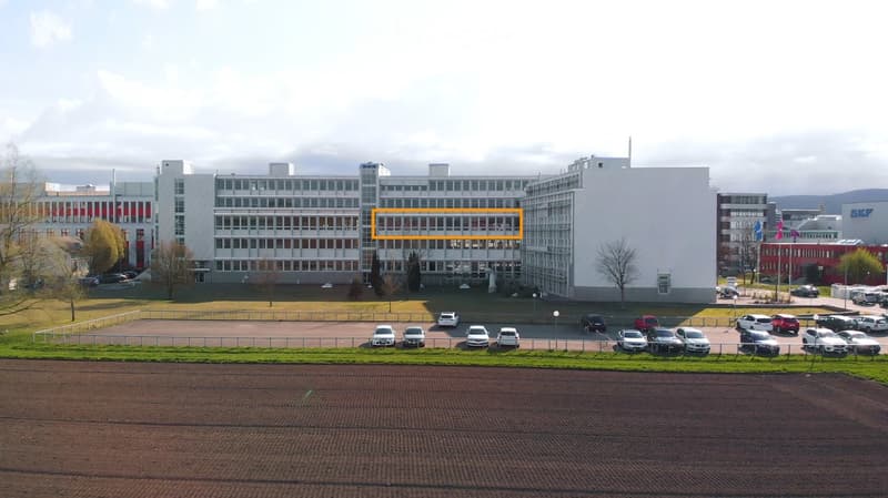 A large, modern multi-story building with a white facade and numerous windows, surrounded by a parking lot with several cars and a grassy area.
