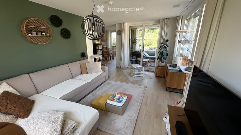 Modern living room with a white sectional sofa, coffee table, and a dining area visible through an open doorway. The space features light wood flooring, large windows, and a balcony accessible through glass doors.