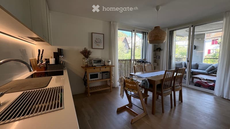 A modern, well-lit kitchen and dining area with wooden flooring, a sink, microwave, toaster, dining table, chairs, and a sofa visible through a large window. The space features a balloon lamp and a picture frame on the wall.