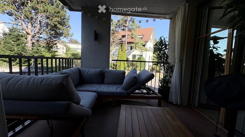 Balcony with wooden decking, grey sectional sofa, small wooden table, potted plant, and string lights.