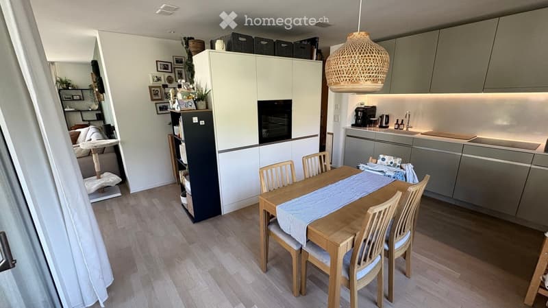 Open-plan living and dining area with modern kitchen, dining table with chairs, pendant light, and shelves with decorative items.