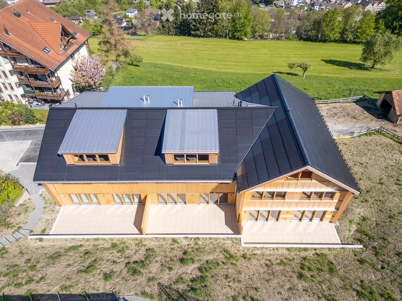 A modern two-story house with a dark grey metal roof, solar panels, and large wooden-framed windows. The house features a spacious patio area with a tiled floor and is surrounded by a green field and trees.