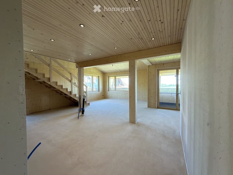 Empty room with wooden ceiling, concrete walls, and a staircase leading to another level. The floor is unfinished concrete, and there are large windows and a glass door leading to an outdoor area.